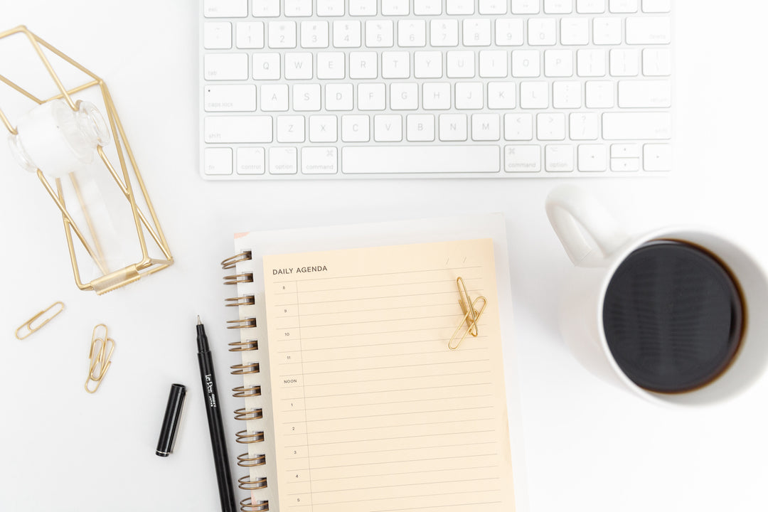 top view of daily agenda book on white desk with coffee mug and apple keyboard